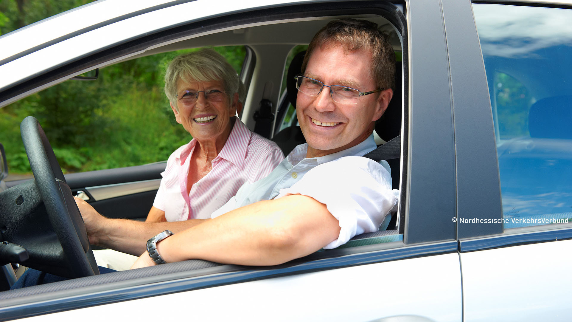 Zwei Personen in einem Auto, die durch das Fenster in die Kamera lächeln.